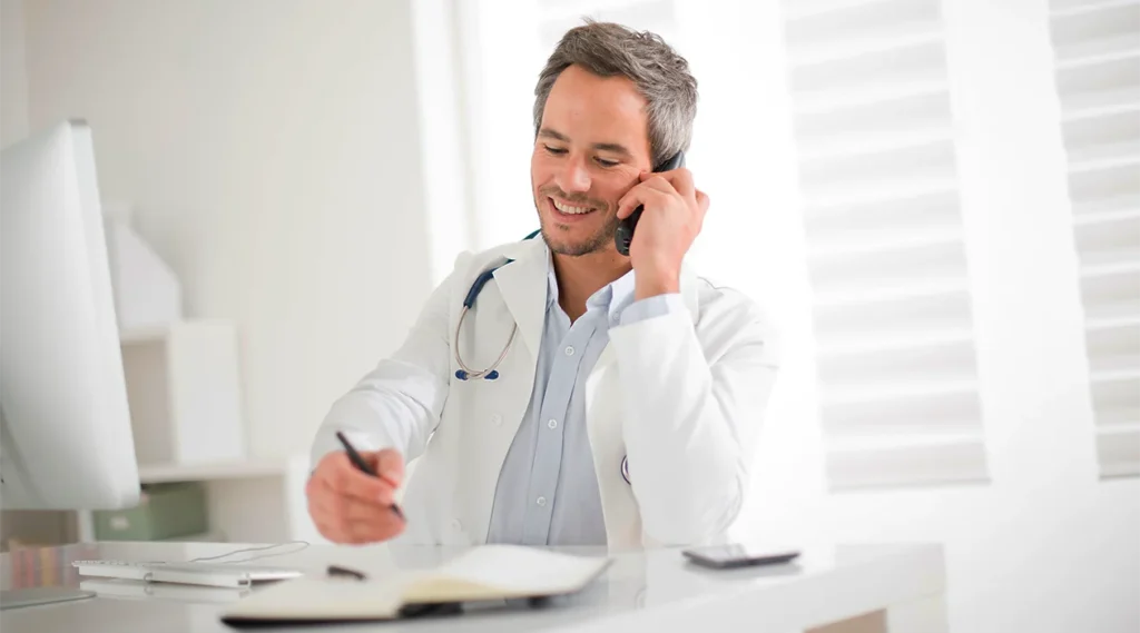 Ein lächelnder Arzt spricht am Telefon, während er an einem Schreibtisch sitzt